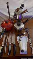Photo showing the entire lot including Le Creuset pot, fondue set, knives, and kitchen utensils arranged on table.