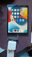 Front view of Apple iPad powered on with home screen apps visible, charging block and cable shown.