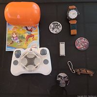 Full lot image showing mini drone with controller, iPod Shuffle, Timex wrist watch, and six collectible pogs on black surface with orange case and cartoon paper underneath