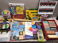 Photo showing an assortment of art supplies including packaged Crayola markers, oil pastels, colored chalk, sidewalk chalk paint bucket, and various art books and craft kits.