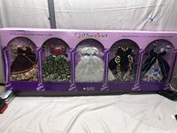 Set of five classic doll dresses with matching jewelry displayed in original purple box, clear windows showing each outfit and accessories.