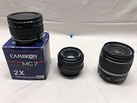Three camera lenses displayed on a white background: Cambron MC7 2X Tele Converter on box, Minolta manual focus lens, Canon EF-S 18-55mm zoom lens.