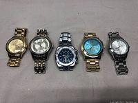 Photo showing all five men’s wrist watches in the lot laid out on a surface, displaying front faces and bands, with varying metal tones and dial colors.