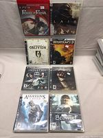 Eight PS3 game cases standing upright, arranged in two columns of four. Titles visible: Prince of Persia, Lair, Oblivion, Resistance 2, Injustice, Assassin's Creed, Beowulf.