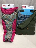 Two dog winter coats side by side: a pink quilted parka with plaid chest panel and a green coat with plaid lining and toggles.