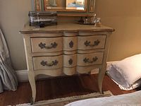 Front view of the decorative cream-colored wood dresser with two drawers and ornate metal pulls, showing its curved design and top surface.