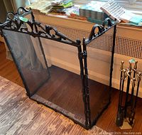 Front angle view of three-sided fire screen showing decorative cast aluminum top and black mesh panels placed on wood floor near window.