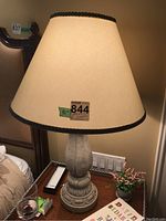 Full view of one lamp showing beige fabric shade with dark trim and carved composite base on wooden side table.