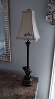 Full view of one table lamp showing fabric shade with bead fringe and metal-ceramic figural base on marble surface.