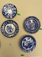 Four blue and white decorative plates on a yellow wall with measurement tags from 8 to 9.5 inches, two plates have intricate scenic and floral designs, two have patterned borders