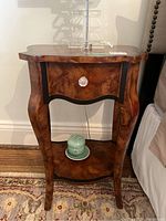 Front view of composite side table with faux wood veneer, decorative faux drawer with crystal-like knob, and lower shelf holding small ceramic container with lid and plate.