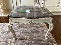 Angle view of the wooden footstool showing carved legs and plaid upholstered top with brass nailhead trim.