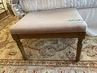 Front view showing bench with wooden legs and upholstered top in beige fabric, placed on decorative carpet.