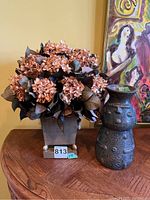 Full display of ceramic vase with face and square metal planter with dried light brown flowers on wooden surface with painting in background.