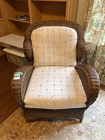 Front view of brown wicker arm chair with off-white seat and back cushions on a patterned rug near wooden furniture.