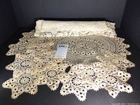 Rolled and displayed large off-white circular lace tablecloth with scalloped edges and detailed floral/lace pattern visible.