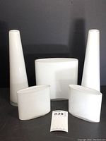 Frontal view of three iitala Tapio Wirkkala Ovalis white art glass vases with two taller Tarnoweic cylindrical vases behind on black background.