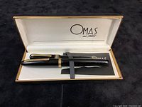 Two black Omas pens inside open cream-colored Omas box with logo, displayed with manual.