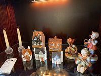 Wide shot of 3 Hummel figurines and 4 British cottage ceramic figurines with 2 glass candle holders in background