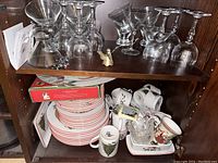 Wooden shelf with stacked Christmas china plates, mugs, a Christmas-themed box, small decorative trays, and a small figurine. Also visible glassware on upper shelf.
