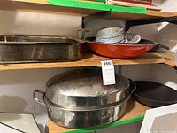 Photo of cookware set on wooden shelf including turkey pan, roasting pan, cast iron pan, orange wok with disposable pans inside.