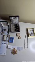 Photo of assorted jewelry including rings in a ring box, earrings in two boxes, a brooch with label, and various charms and necklaces laid out on white surface.
