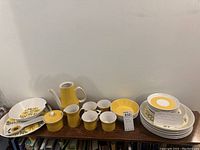 Wide view of most of the ceramic pieces including tea pot without lid, creamer, sugar bowl with lid, mugs, and plates.
