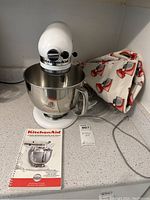 Front view of white mixer with bowl attached, fabric cover, instruction manual and auction tag