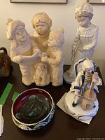 Wide view of the lot showing three cream porcelain statues, the music player figurine, and a decorative bowl placed on a wood surface.