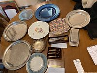 Photo showing multiple silver trays, blue metal tray, ceramic plates, bronze butter dish, and boxed flatware on a wooden table.