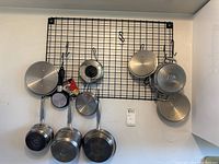 Full view of all pans, lids, and black metal hanging rack mounted on a white wall with hooks holding the cookware.