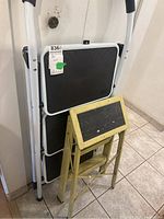 Two step stools are shown standing against a wall: a large black and silver folding stool and smaller yellow metal stool.