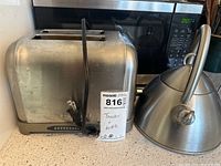 Photo showing stainless steel two-slot toaster and stove kettle on countertop with microwave in background.