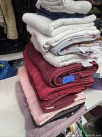 Stack of 12 towels in various colors including white, navy, pink, maroon, purple, beige. One towel with striped pattern identified as beach towel.