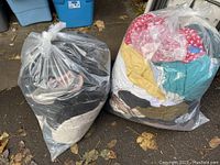 Two large clear plastic bags filled with assorted his and hers clothing and shoes, positioned on the ground outside.