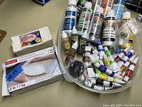 Lot of bottles and tubes of acrylic paint, resin, modeling clay, and war paint tubes shown in container and next to a large package of white air dry modeling clay.