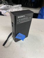 Photo showing vintage Sony AM/FM transistor radio in black plastic body with speaker grille and frequency scale visible, wrist strap attached.