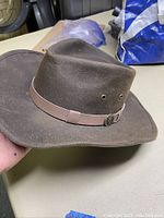 Side angle of a dark brown oilskin hat with a light brown leather hatband and metallic badge.