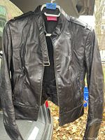 Front view of the black leather ladies jacket hanging on a hanger with zippered pockets, stand collar with buckle, and shoulder epaulets with buttons.