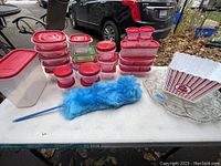 Plastic food storage containers with red lids arranged in stacks on a table, along with a blue feather duster and a white popcorn container holder