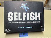 Photo of the Selfish Space Edition card game box, black box with astronaut graphic and game details.