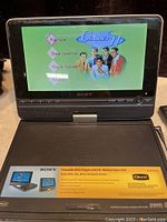 Sony portable DVD player displaying a DVD menu screen on its 9-inch widescreen LCD. The player is black with control buttons and branding visible.