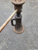 Front view of antique cast iron screw jack resting on asphalt surface with rusted metal handle inserted.