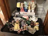 Avon collectible perfume figurines and decorative items arranged on shelf and black storage bin lid, showing multiple figurines including women in vintage dresses, a man, cherub, and gnome.