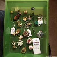 Top down photo of a green tray with assortment of brooches, pins and pendants displayed, showing the quantity and variety of jewelry items.