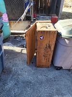 Wooden military crate for Civil Defense First Aid Kit shown standing upright with open door visible, placed outdoors among other containers.