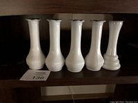 Six milk glass bud vases arranged in a row on a wooden shelf. Five vases have ribbed vertical lines and flared rims; one has a smooth surface and rounded base.