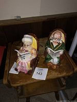 Two ceramic retirement saving bank figurines of an elderly man and woman sitting in armchairs, viewed from the front.