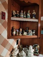 Porcelain figurines arranged on two wooden shelves with lace trim, top shelf has seven human character figurines, middle shelf has eight figurines including animals. Bottom counter shelf shows a white porcelain gravy boat, matching teapot, and snowman figurine.