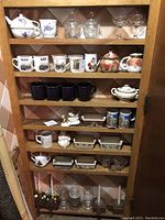 View of wooden shelf with multiple ceramic mugs, ramekins, salt and pepper shakers, decorative boxes, and glassware items.